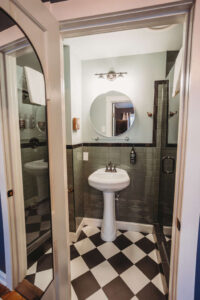 A small bathroom featuring a pedestal sink, round mirror, and glass shower door.