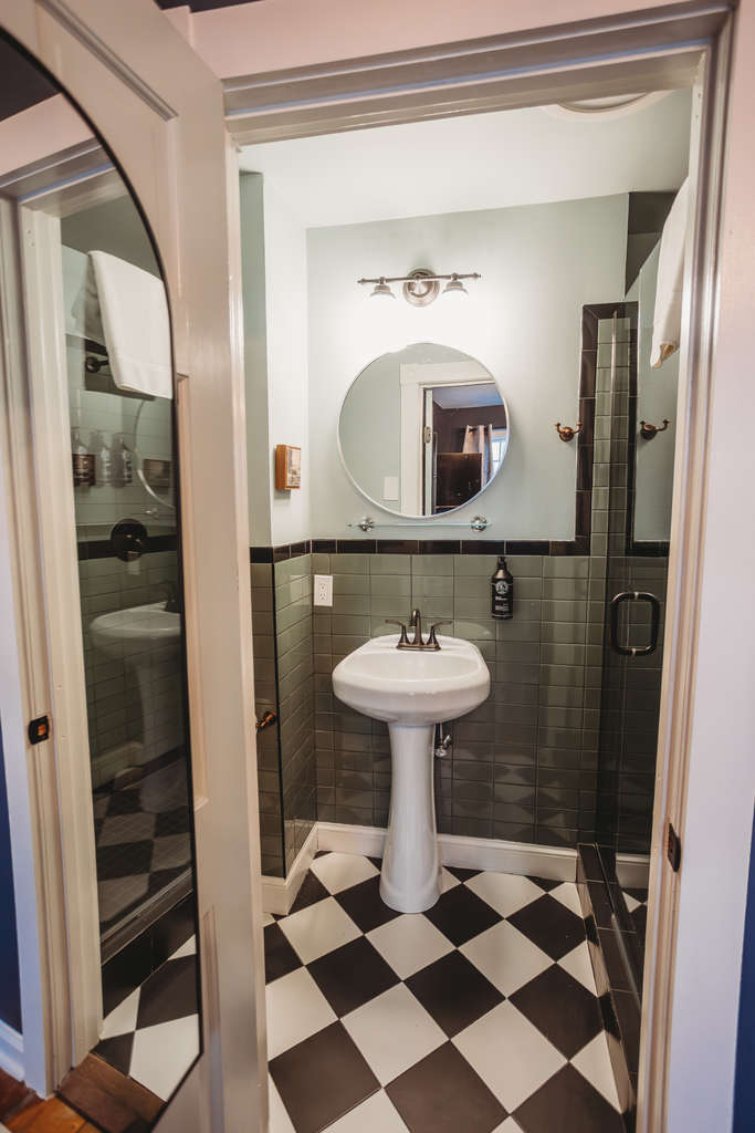 A small bathroom featuring a pedestal sink, round mirror, and glass shower door.