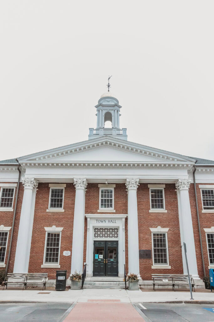 A historic brick town hall featuring white columns, a central entrance, and a clock tower on top.