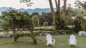 Two white Adirondack chairs on a lush green lawn with trees and mountains in the background.