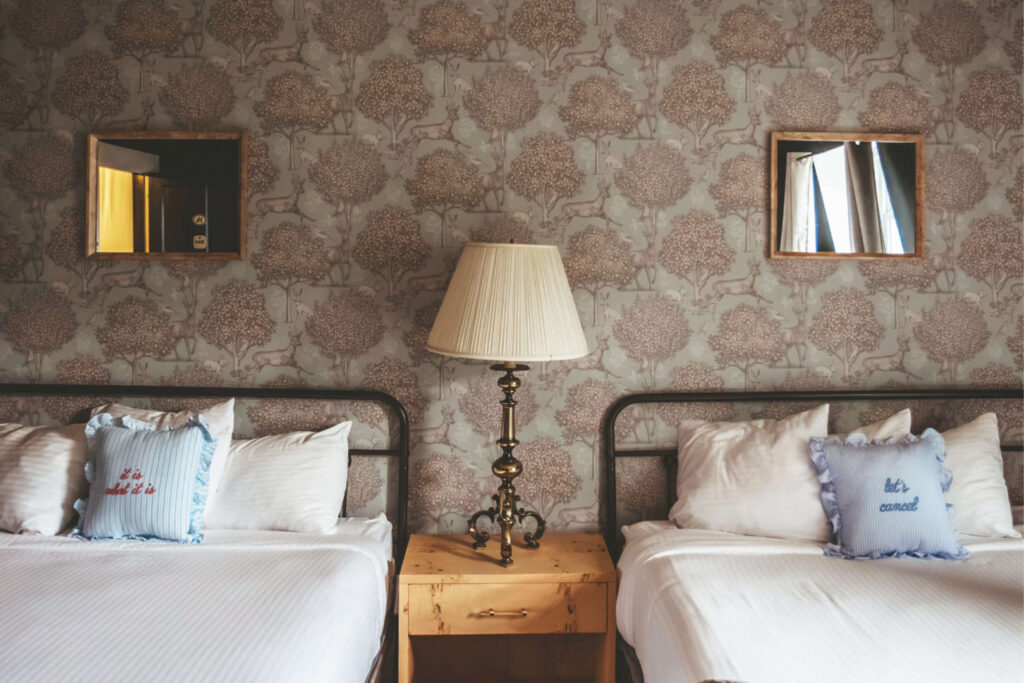Twin beds with decorative pillows, vintage wallpaper, and a bedside lamp.