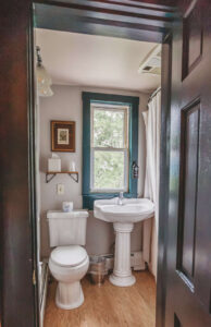Small bathroom with vintage pedestal sink, toilet, and window with curtains.