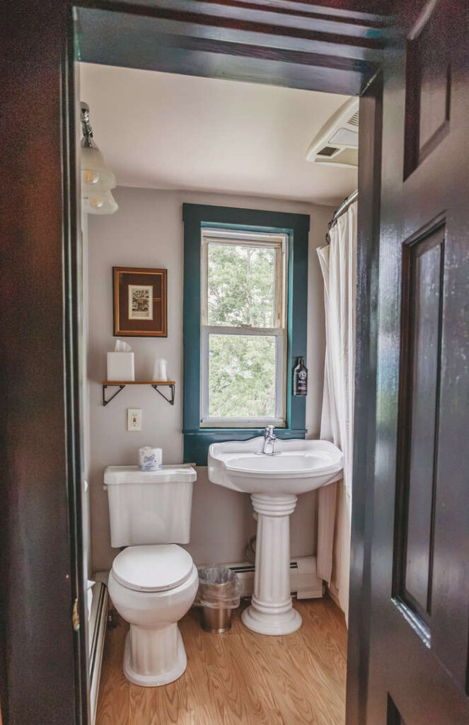 Small bathroom with vintage pedestal sink, toilet, and window with curtains.