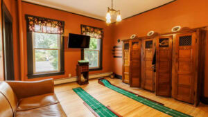 Warm hotel room with wooden lockers, TV, and two windows showing greenery.